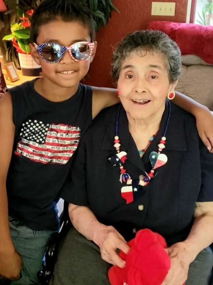 A smiling child in American flag sunglasses and shirt posing with an arm around a seated person wearing a Texas necklace.