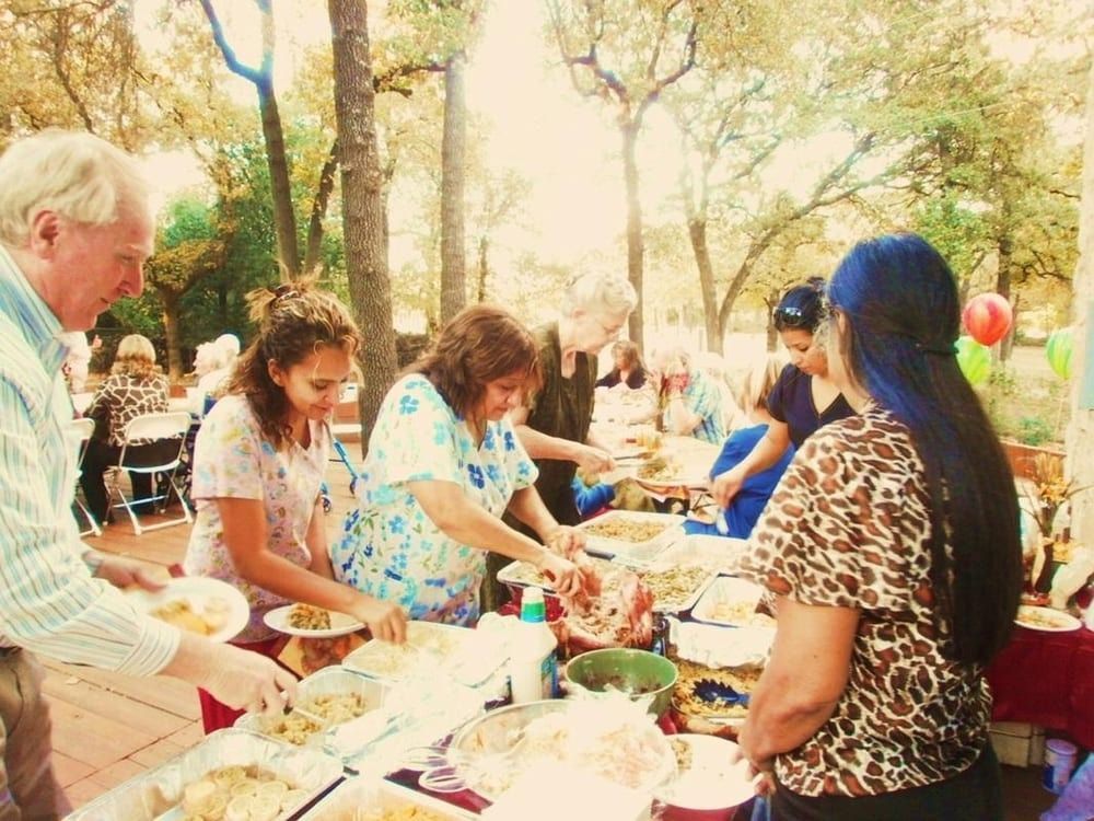 People gather outdoors to serve themselves food from a buffet-style table set up in a wooded area.