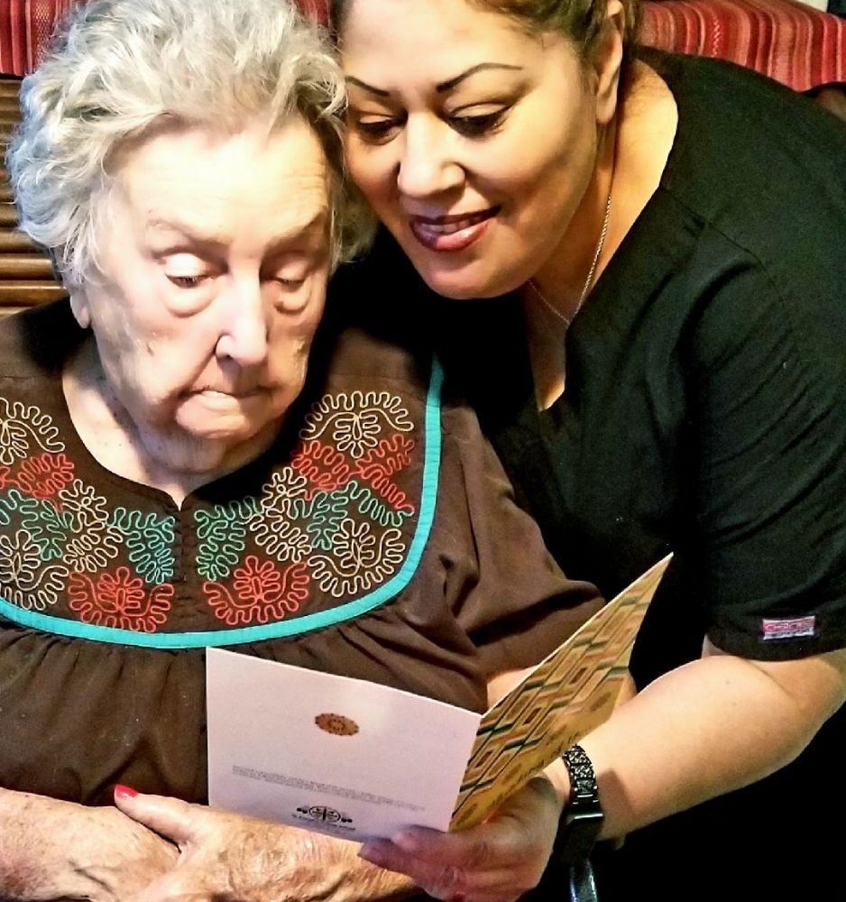 A person in a black scrub top leans over to help an older individual read a greeting card.