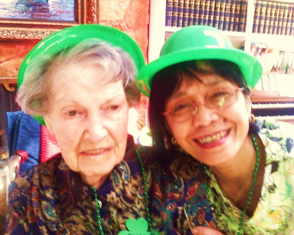 Two people wearing green hats and shamrock necklaces smile side-by-side in a room with a bookshelf.