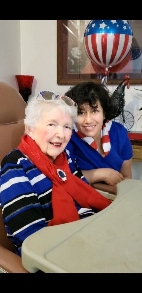 An elderly person and a person in a blue scrub top pose together smiling near a red, white, and blue patriotic balloon.