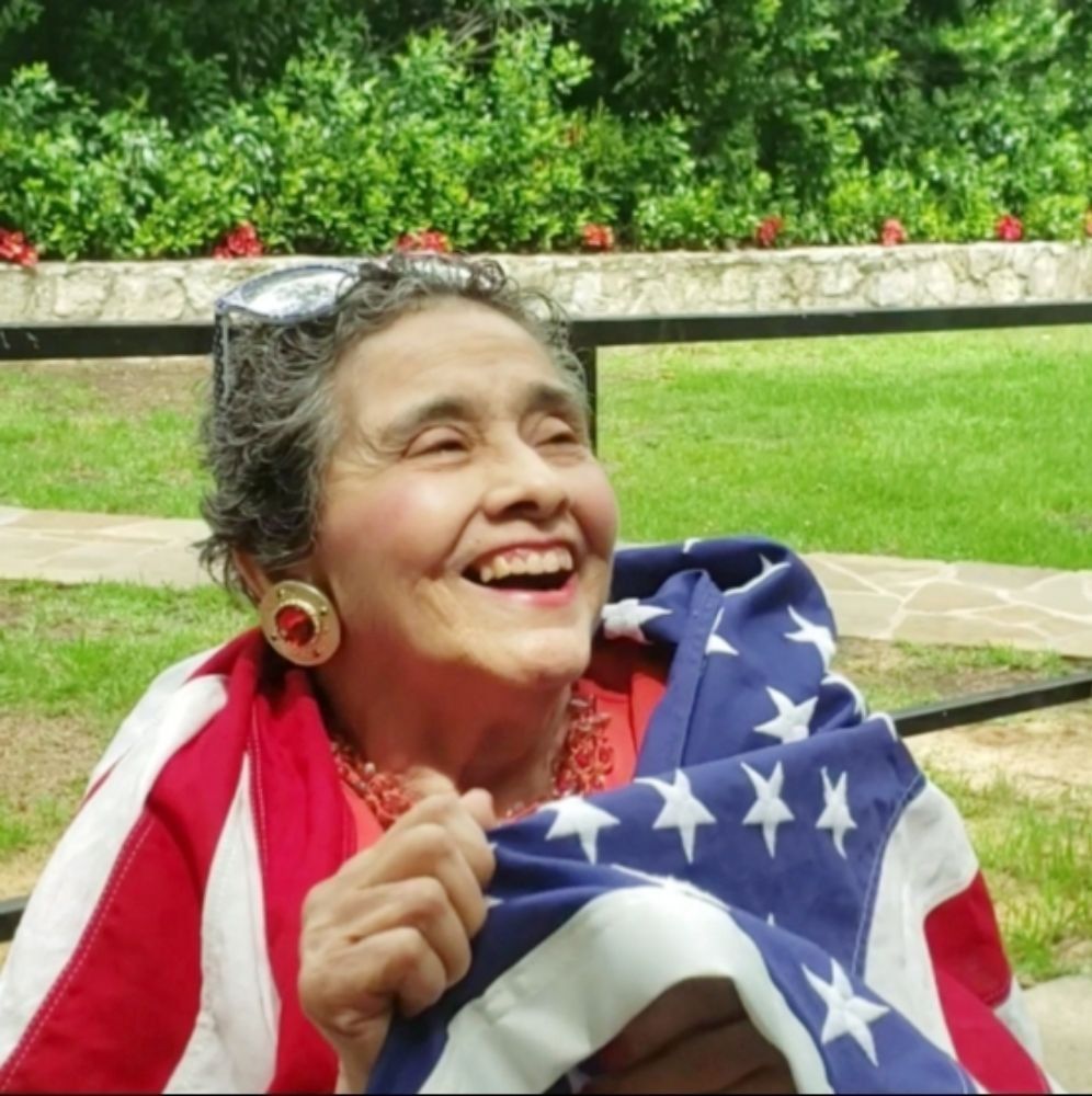 A person smiling outdoors, draped in an American flag with white stars and red and white stripes.