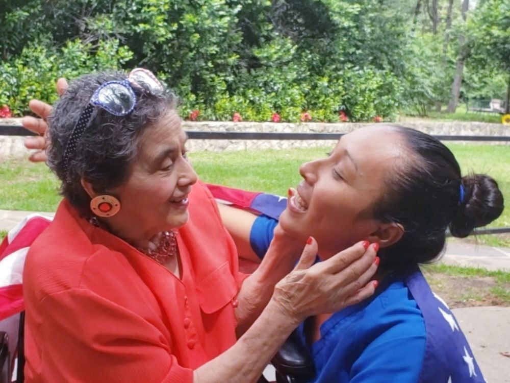 Two people embrace in an outdoor, green setting, one in a red shirt touching the face of the other in a blue shirt.