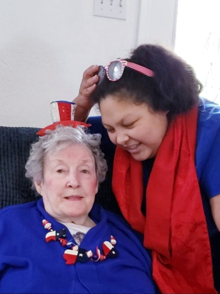 A smiling person wearing a red scarf leans over another person wearing a miniature top hat and a festive necklace.