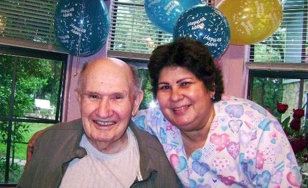 An older person and a woman smiling next to each other in a room decorated with birthday balloons.