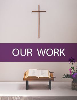 There is a cross on the wall above a bible on a table.