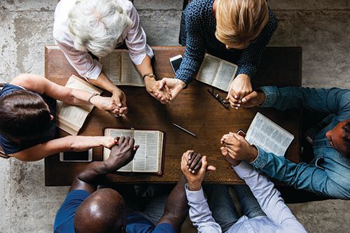 A group of people are sitting around a table with their hands folded in prayer.