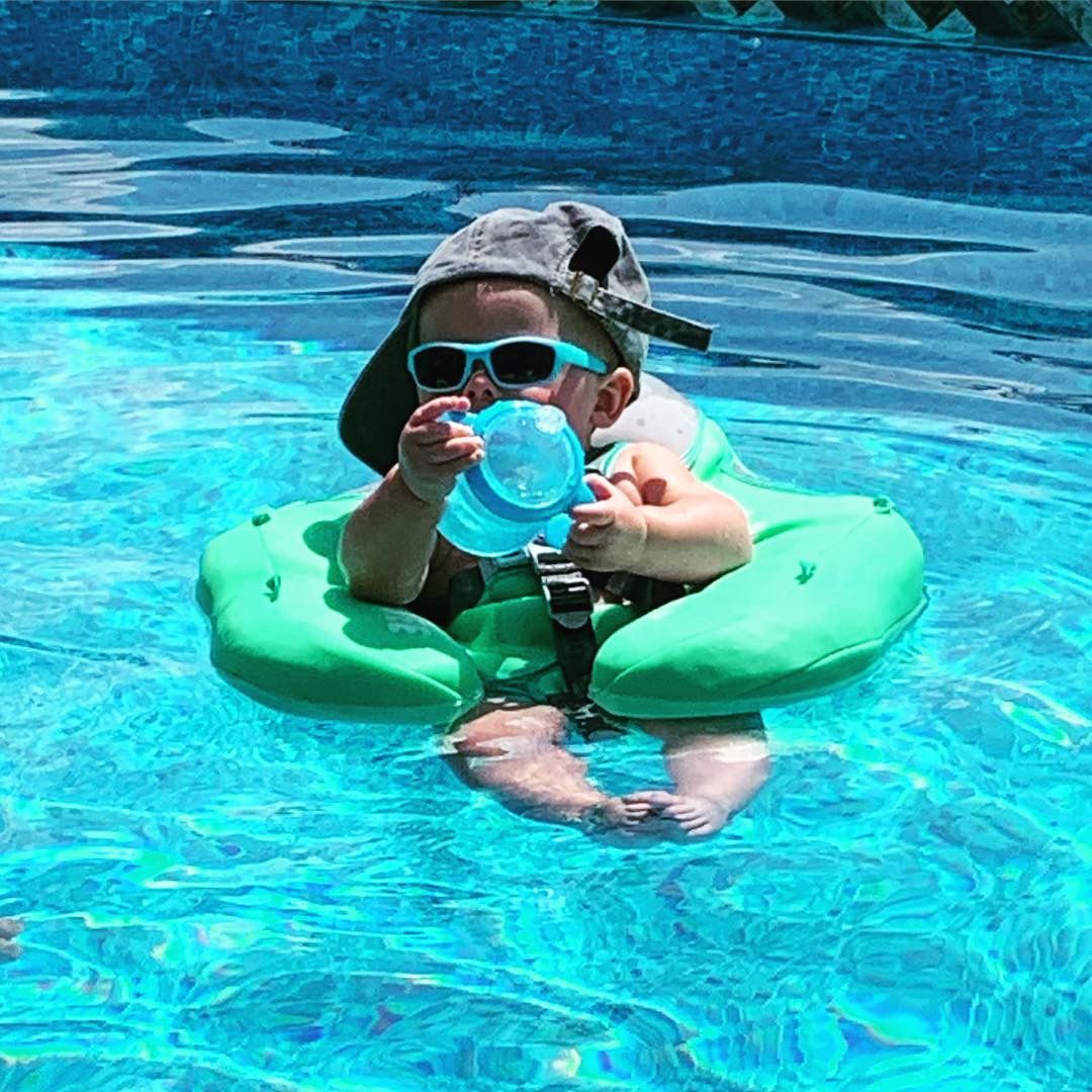 Child in a green float ring in a blue swimming pool, wearing sunglasses and holding a blue cup