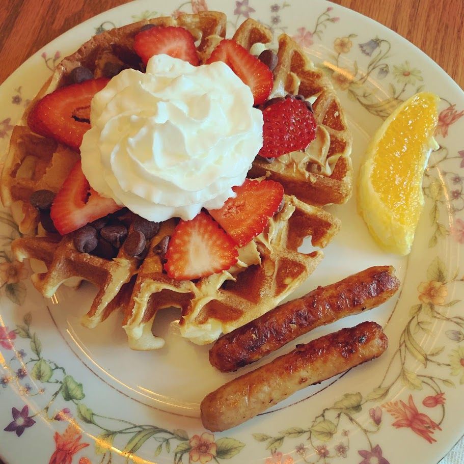 Waffle topped with whipped cream and strawberries, with sausage links and an orange slice on a floral plate.