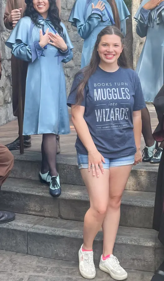 Girl in a blue “Muggles to Wizards” shirt posing on steps at a theme park, with people in robes behind her