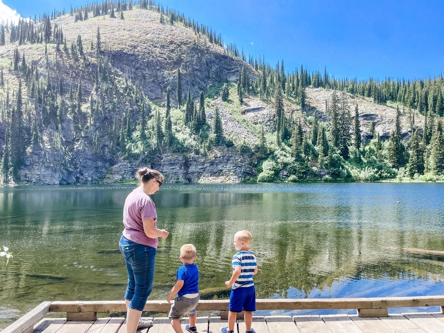Three people stand on a dock by a calm mountain lake with forested hills in the background.