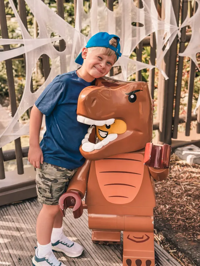 Boy posing beside a brown dinosaur costume in a wooded park setting