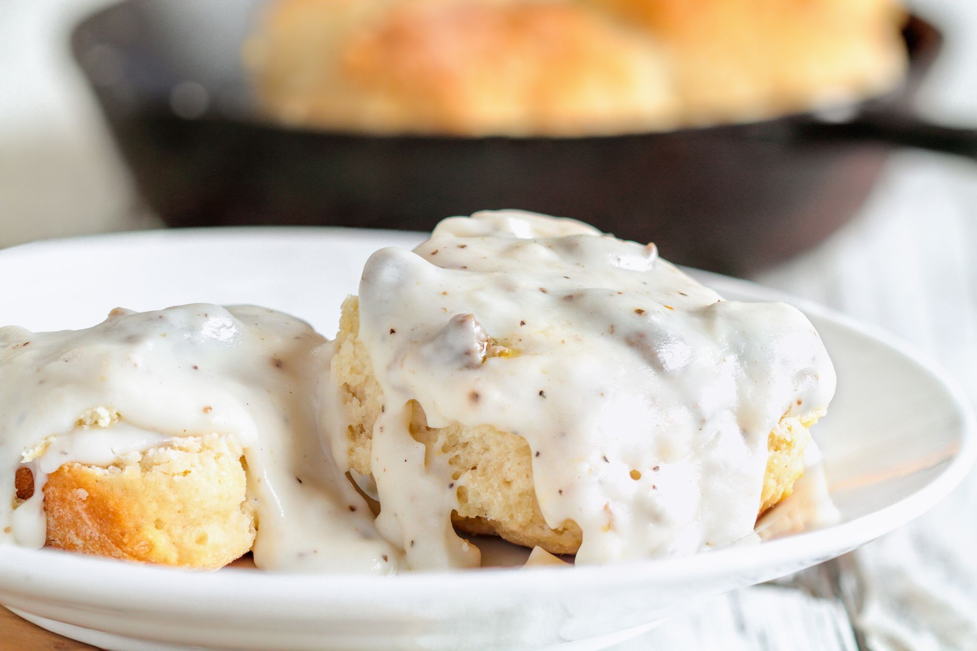 Two golden-brown biscuits on a white plate covered in creamy, peppered sausage gravy with a skillet in the background.