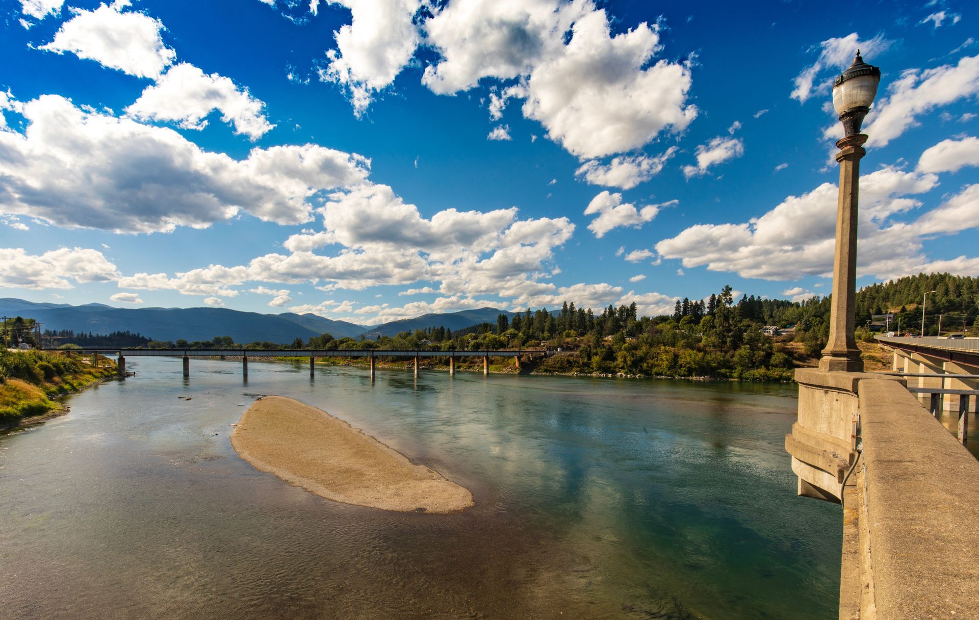 Wide river under a blue sky with clouds, mountains in the distance, and a bridge and lamp post on the right