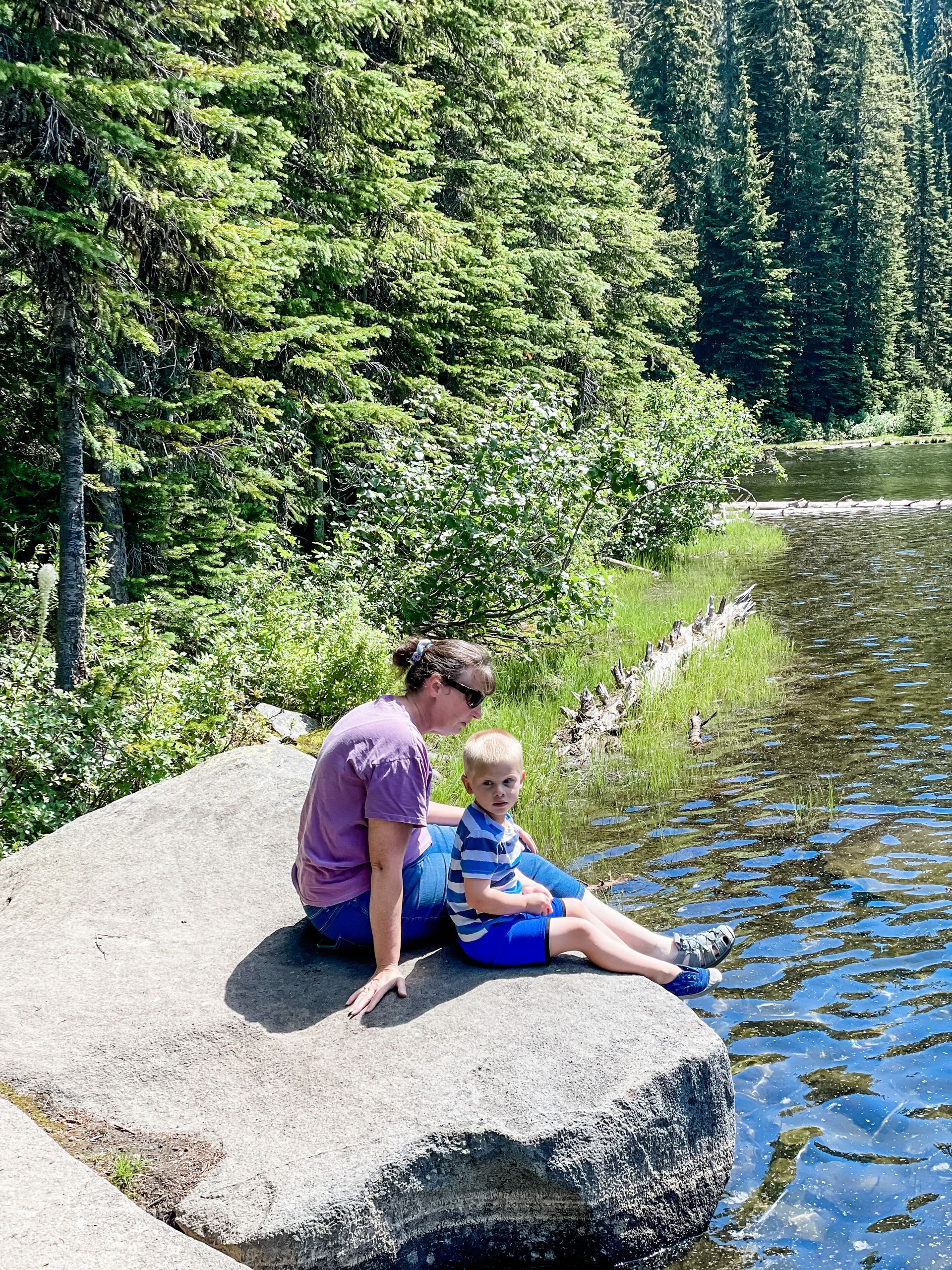 Adult and child sitting on a large rock by a forest lake