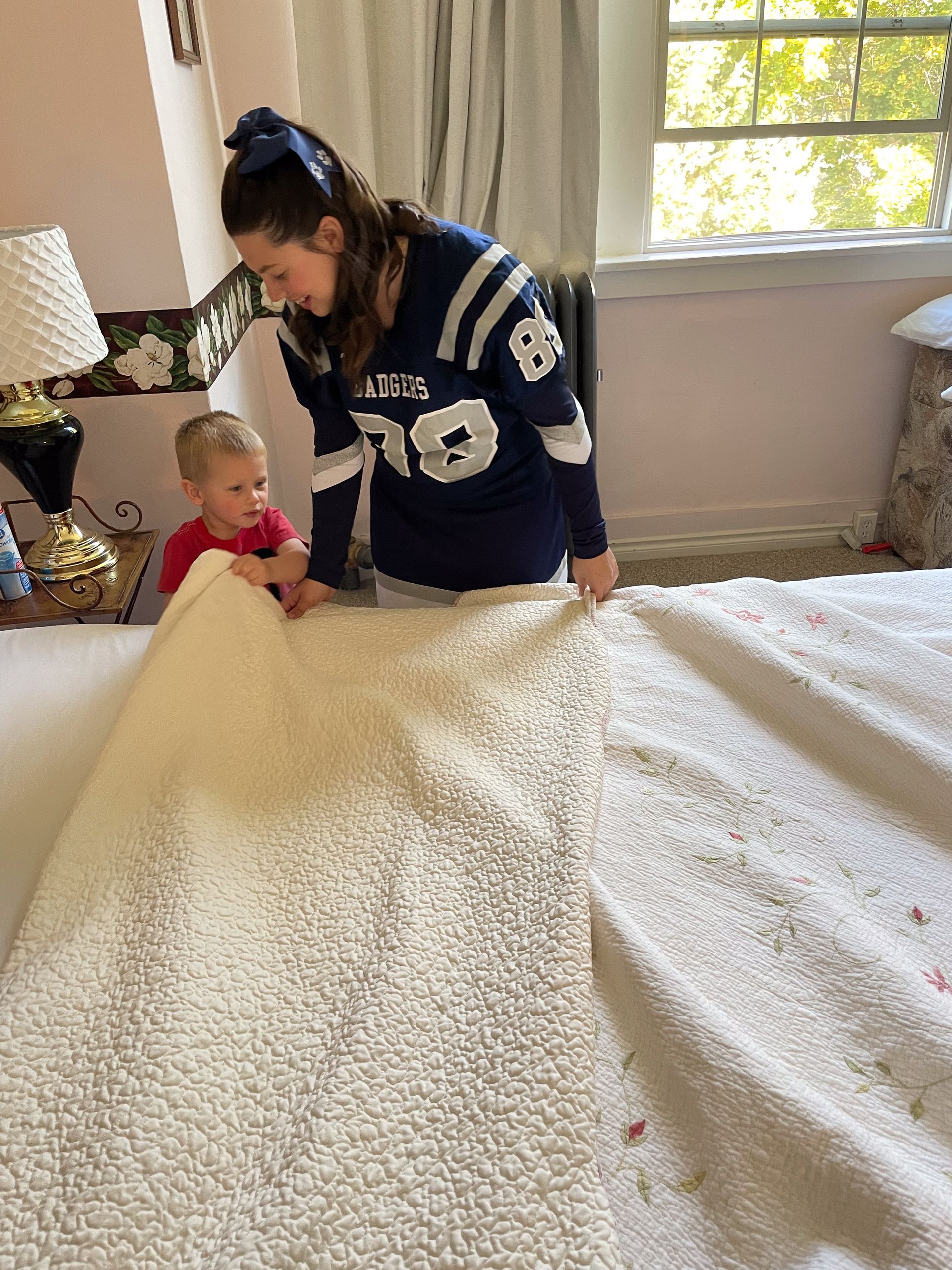 Adult helping toddler under white blanket in sunlit bedroom