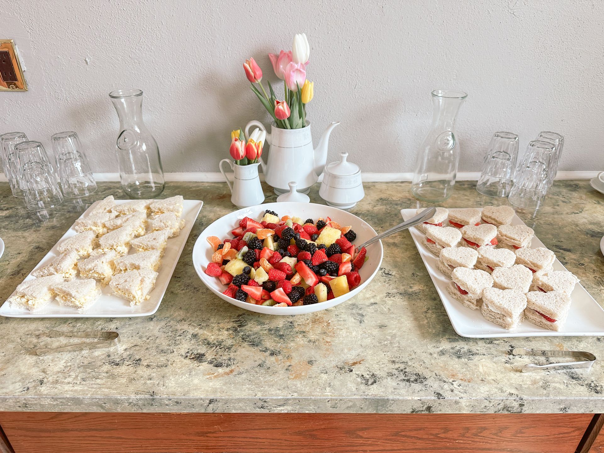 Dessert table with fruit bowl, pastries on platters, and small flower vases on a marble countertop