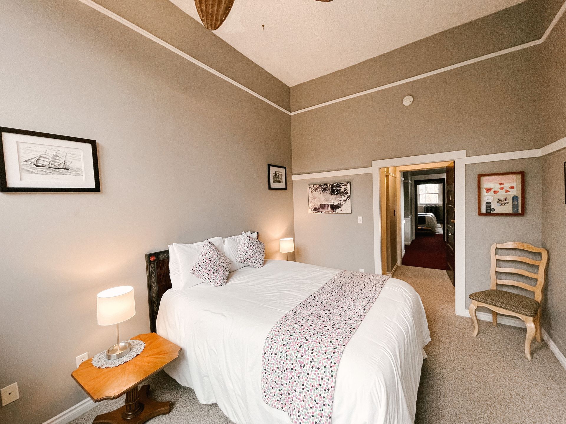 A cozy bedroom with a made bed, a wooden side table with a lamp, a wooden chair, and framed art on neutral-toned walls.