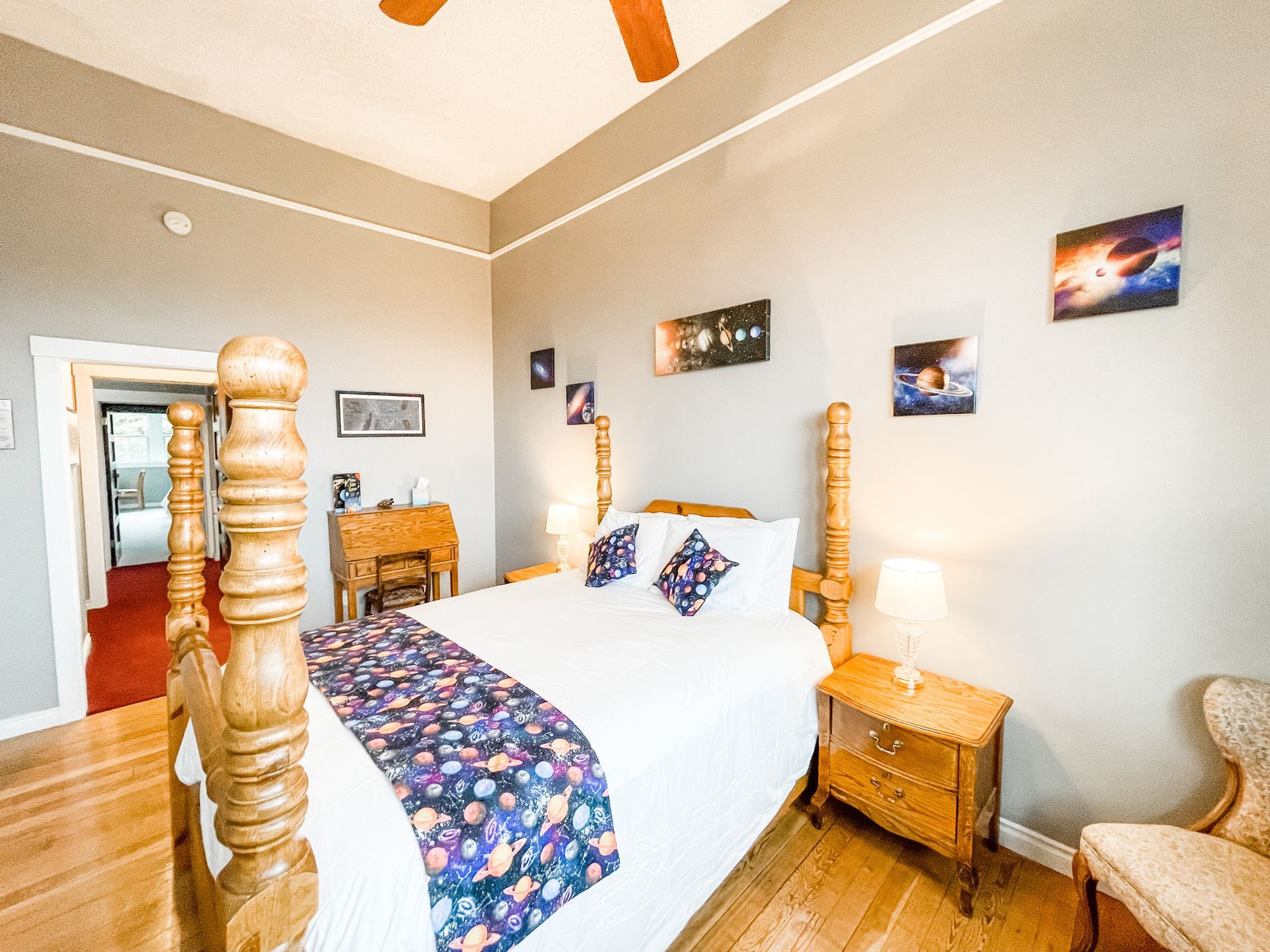 A bedroom with a wooden four-poster bed, space-themed bedding and wall art, two bedside tables, and a wooden desk.