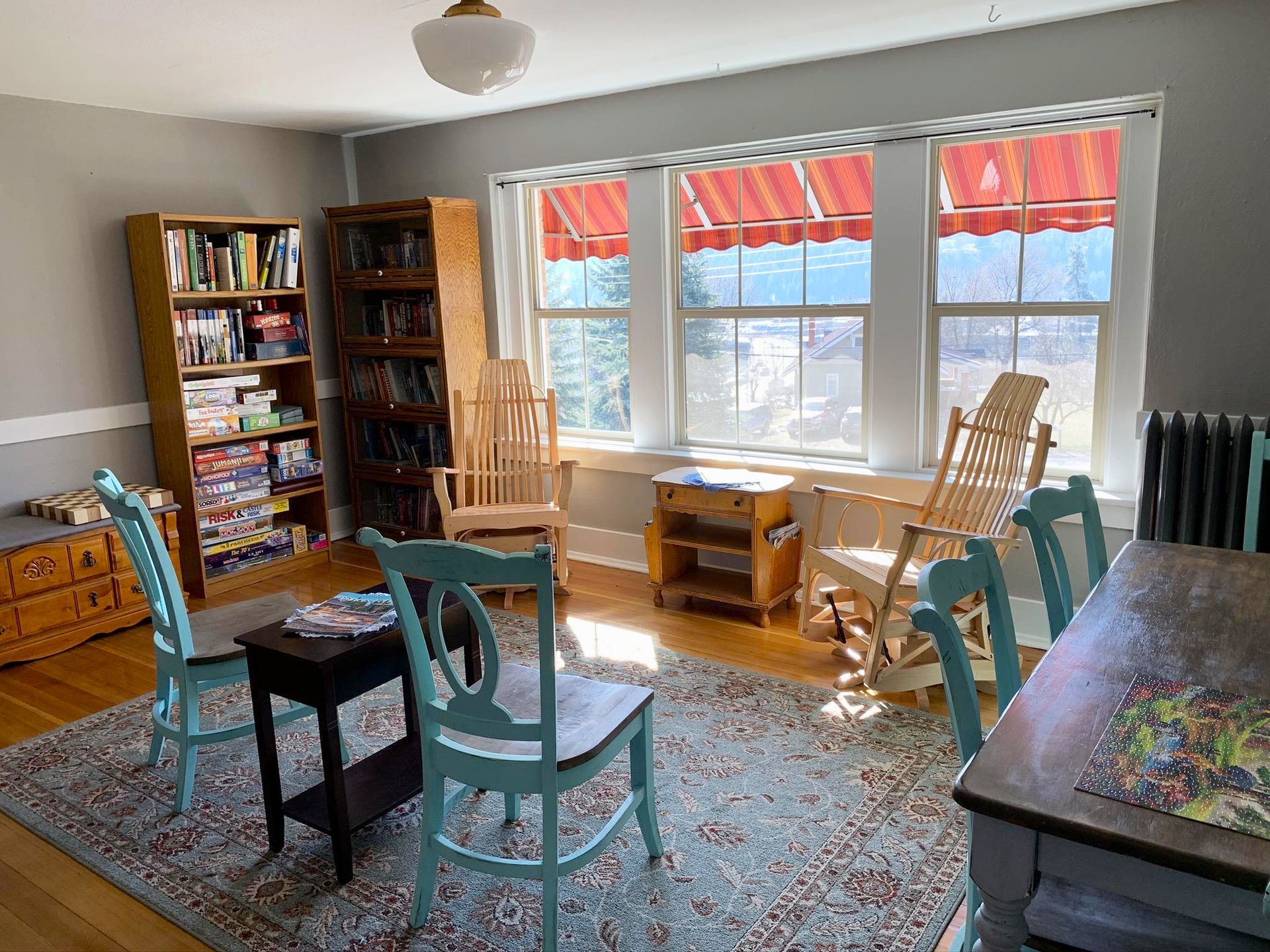 A cozy, sunlit room featuring tall bookshelves, two wooden rocking chairs, and light blue chairs surrounding a small table.