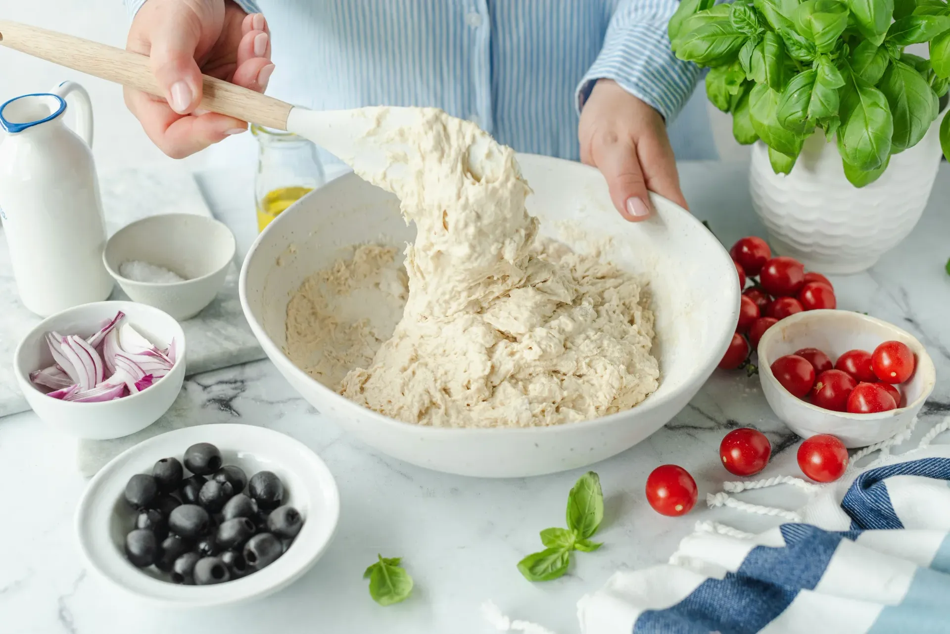 A person mixes pizza dough in a large white bowl, surrounded by ingredients like cherry tomatoes, olives, and basil.