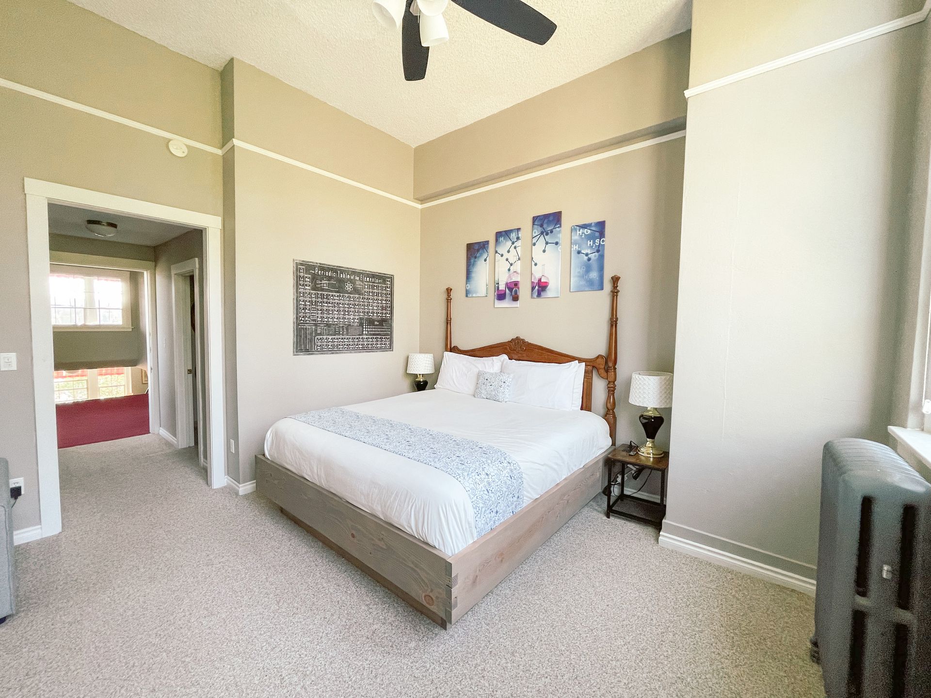 A bedroom with a queen bed, neutral walls, light carpet, four blue abstract art pieces, and an open doorway to another room.