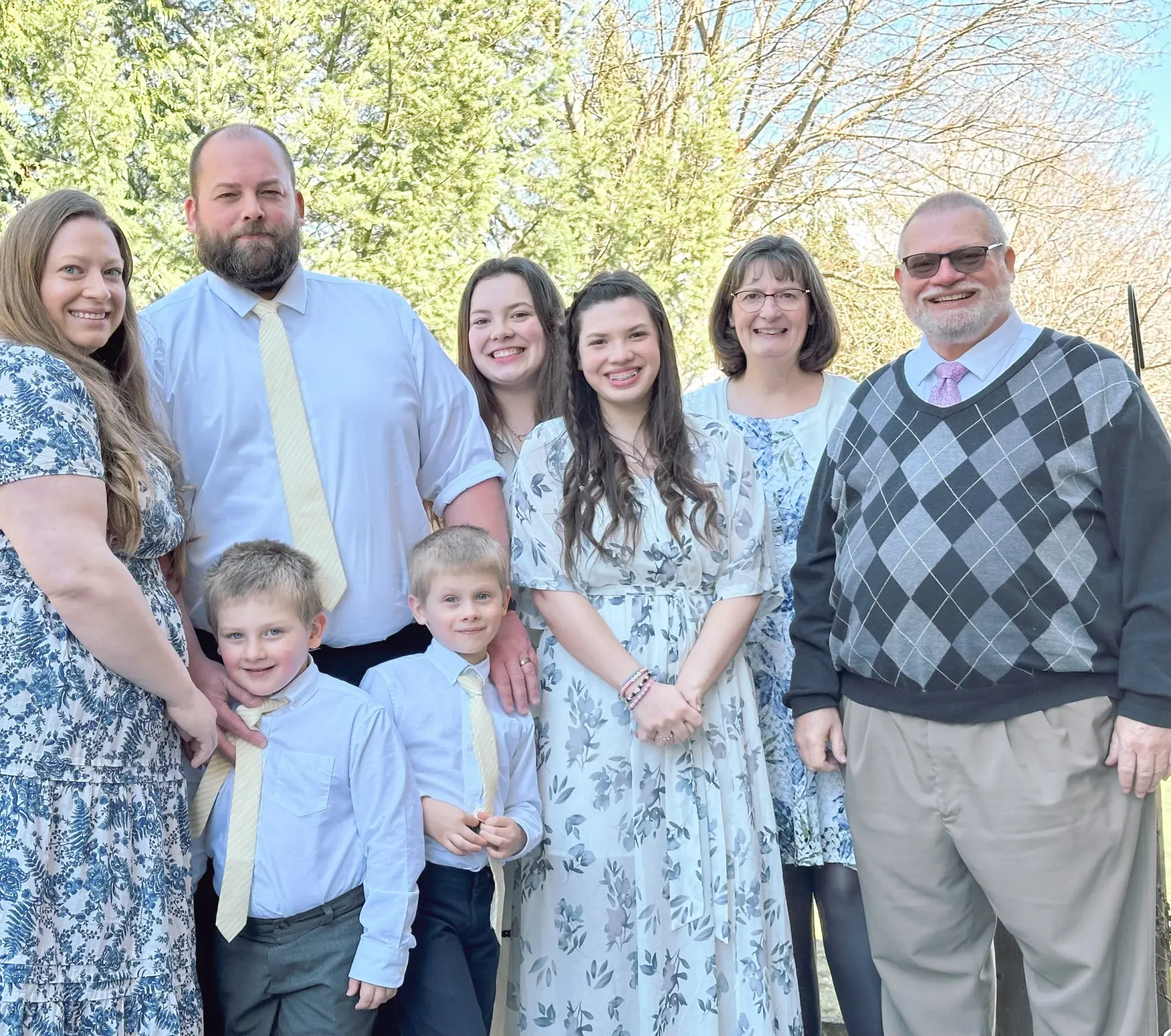A group of eight people stand together outdoors, wearing dress clothes in blue and white, posing for a family portrait.