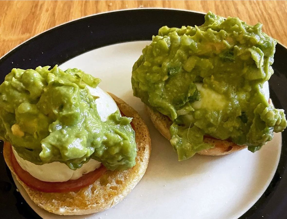 Two English muffin halves topped with sliced tomato, a poached egg, and a thick layer of smashed avocado on a plate.