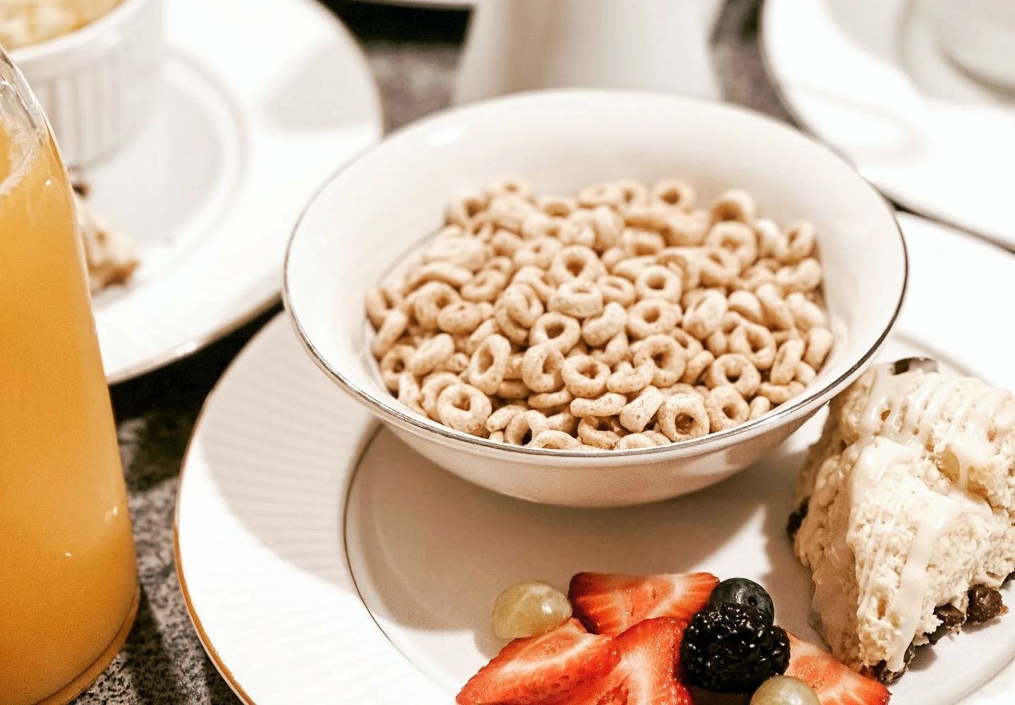 A bowl of circular cereal served on a white plate with strawberries, a blackberry, and a scone, next to a glass of juice.
