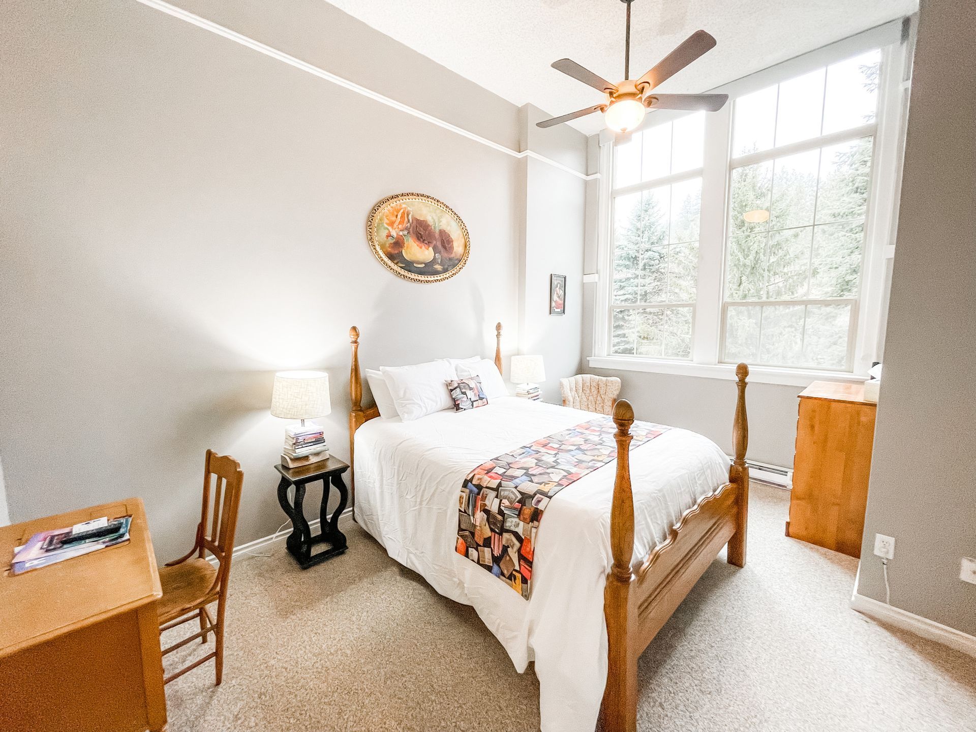 A bedroom with a wooden four-poster bed, a desk and chair, bedside table with lamp, and large windows with natural light.