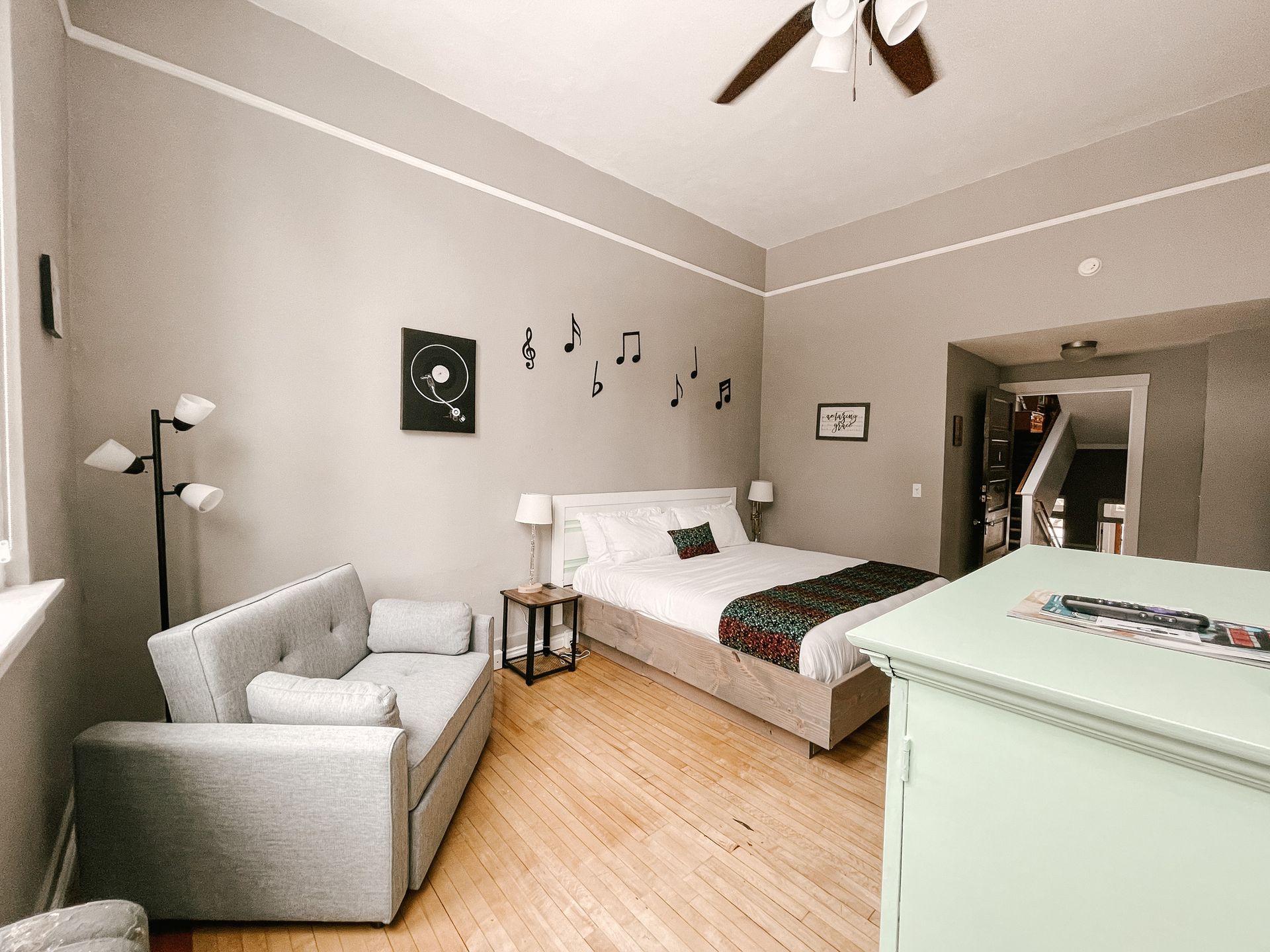 A bedroom with light gray walls, a light gray sofa, a double bed, wooden floors, and wall-mounted musical note decor.