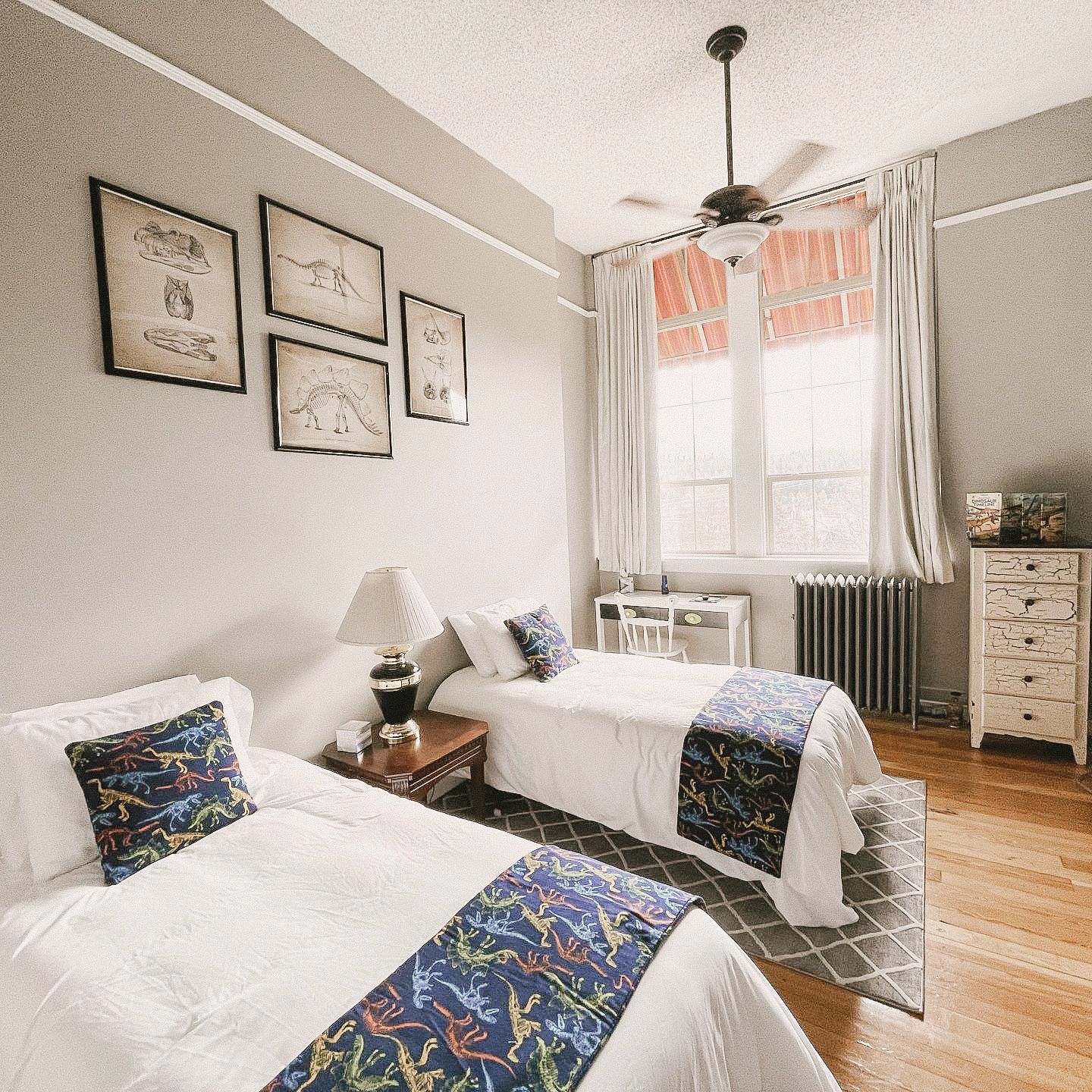 A bright bedroom with two twin beds featuring dinosaur-print pillows, a lamp, wall art, and a large window with curtains.