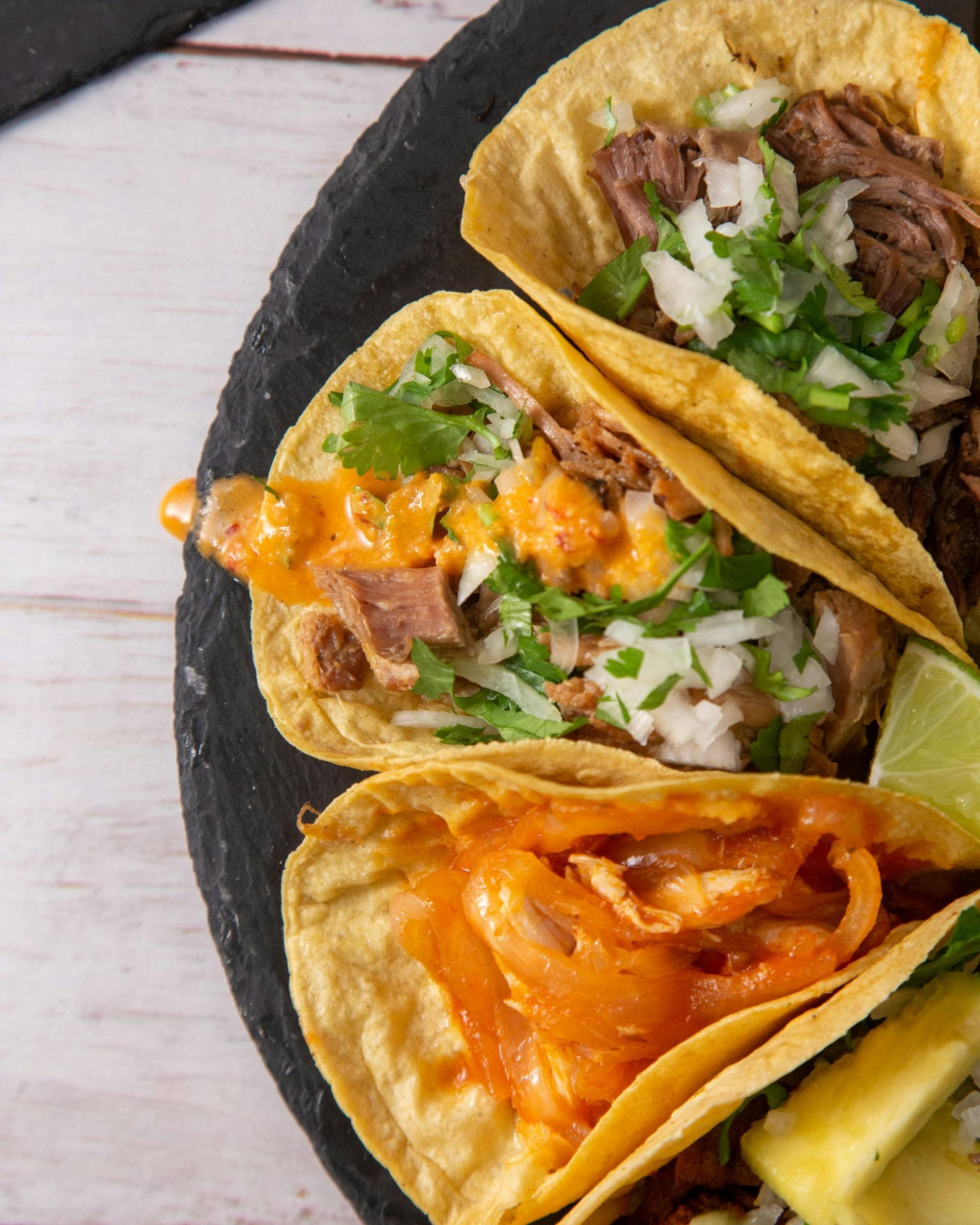 A close-up of three corn tortillas filled with seasoned meat, onions, cilantro, and cheese on a black stone plate.