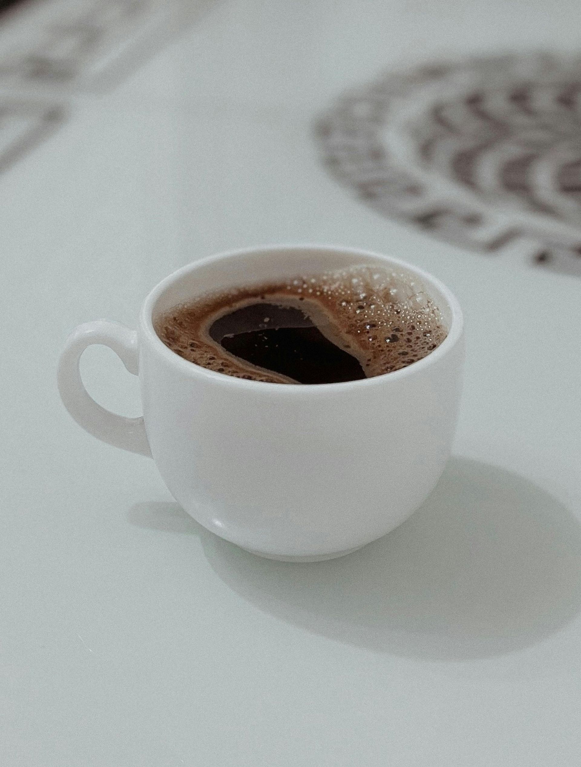 A white cup filled with dark coffee sits on a white surface with a patterned design in the background.