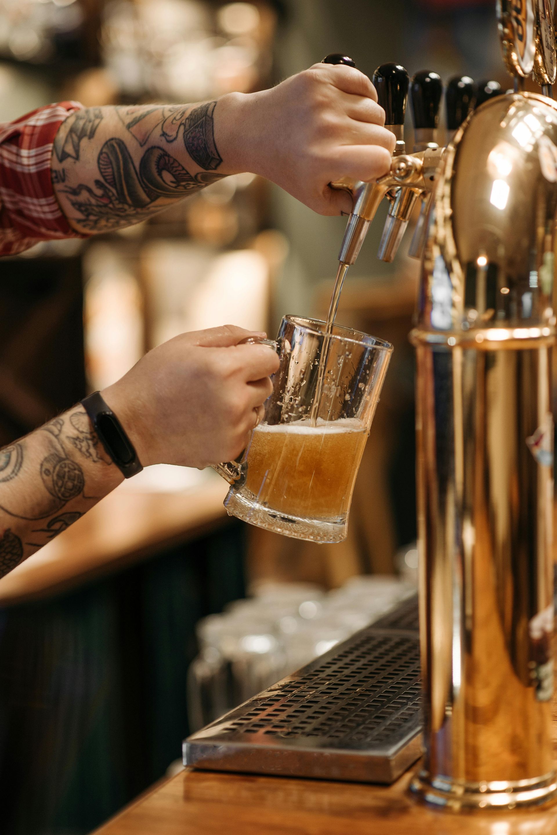 A tattooed person pours beer from a polished golden tap into a glass mug at a bar.