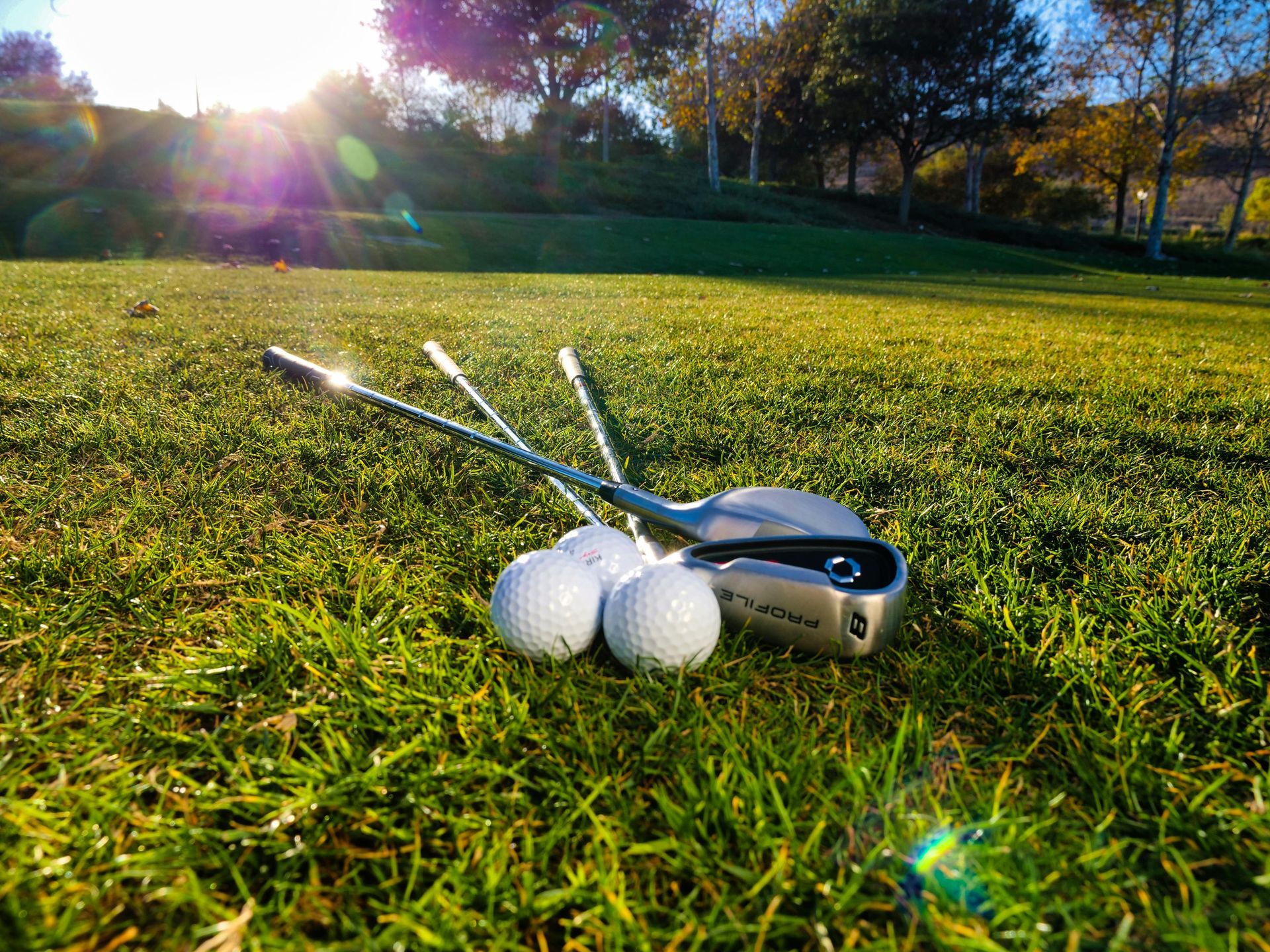 Three white golf balls and two golf irons rest on a grassy green golf course under bright, golden sunlight.