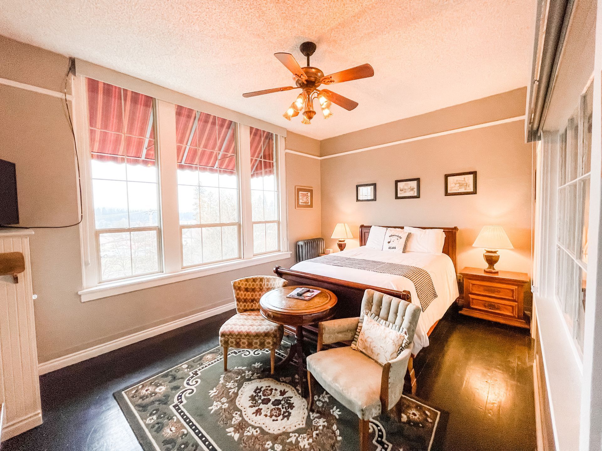 A bedroom with a queen bed, two armchairs, a small round table, a patterned rug, and a ceiling fan under warm lighting.