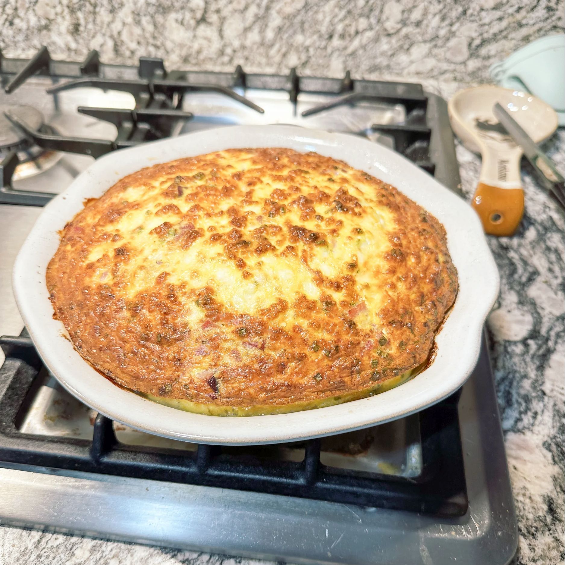 A freshly baked golden-brown quiche in a white scalloped pie dish, sitting on a kitchen stovetop.