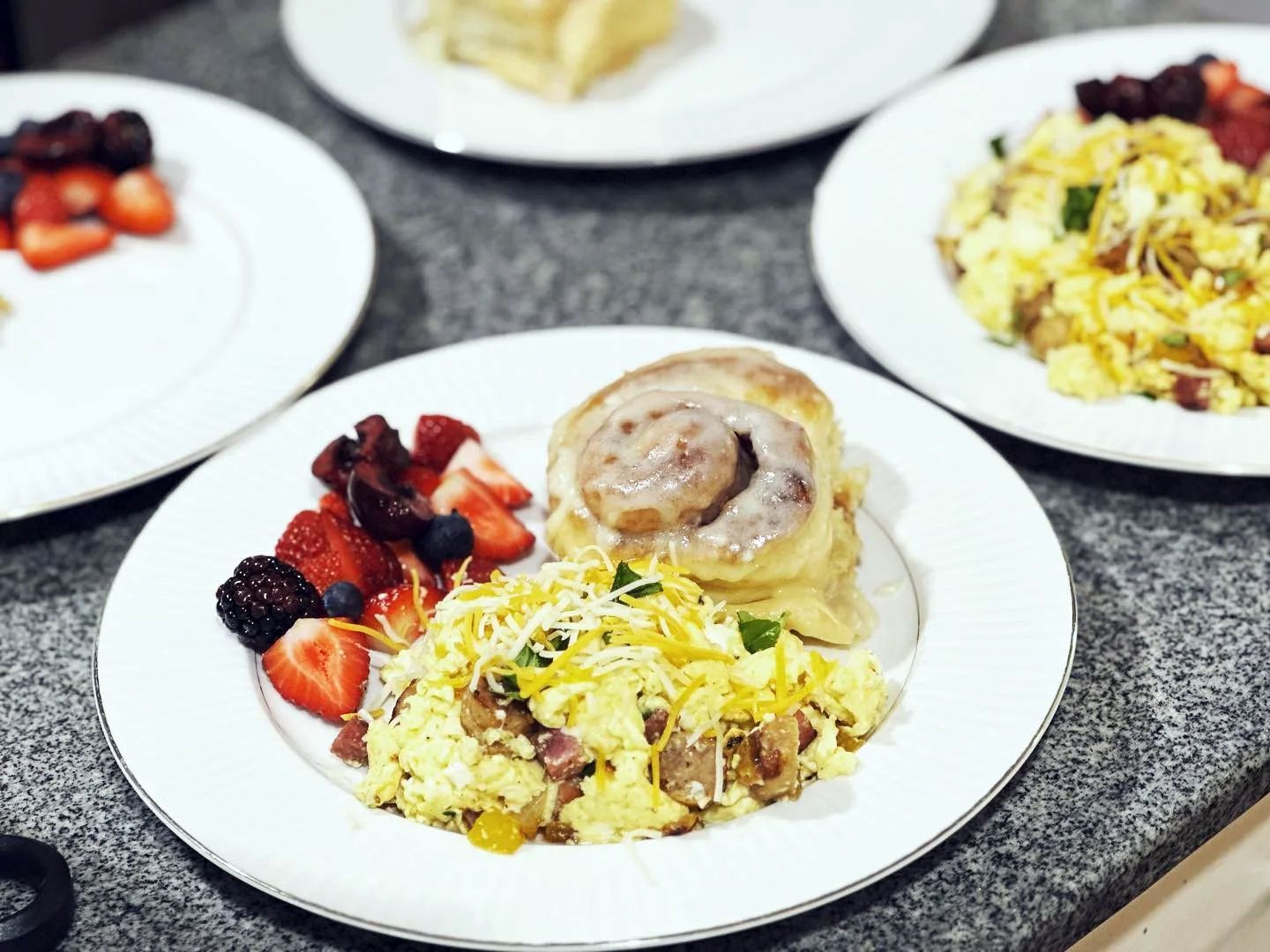 Three plates of breakfast, each with scrambled eggs, a cinnamon roll, and fresh berries, sit on a gray countertop.