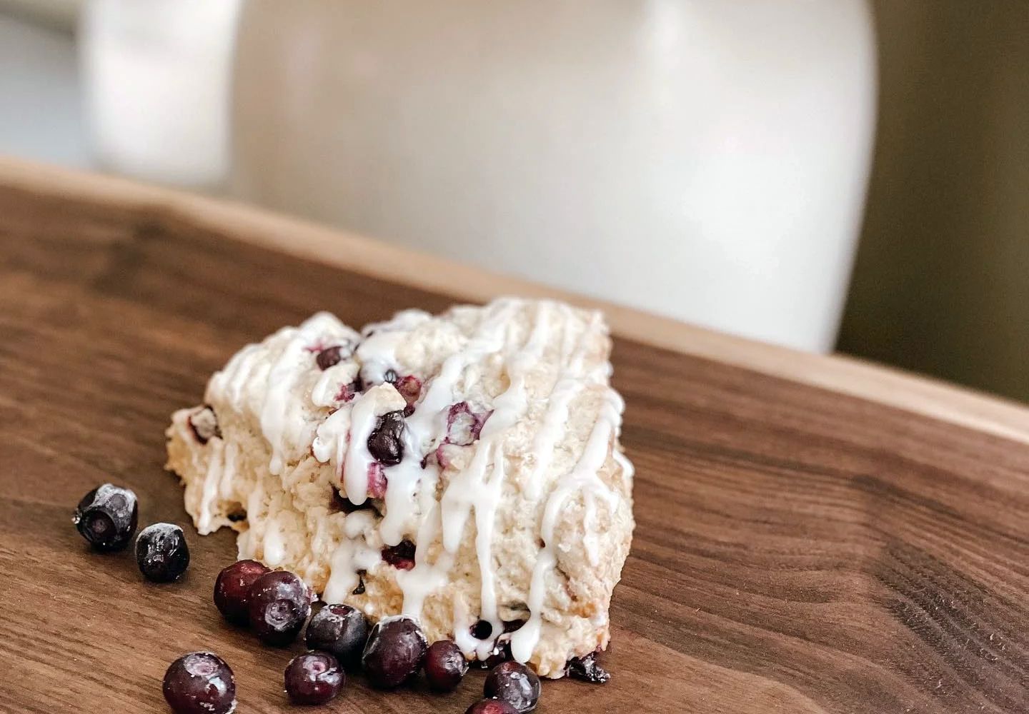 A triangular blueberry scone with white icing drizzle sits on a wooden board, surrounded by loose blueberries.