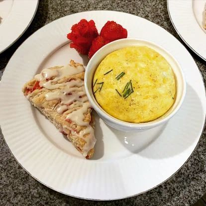 A plate containing a slice of iced pastry, a round baked egg dish topped with chives, and fresh strawberries.