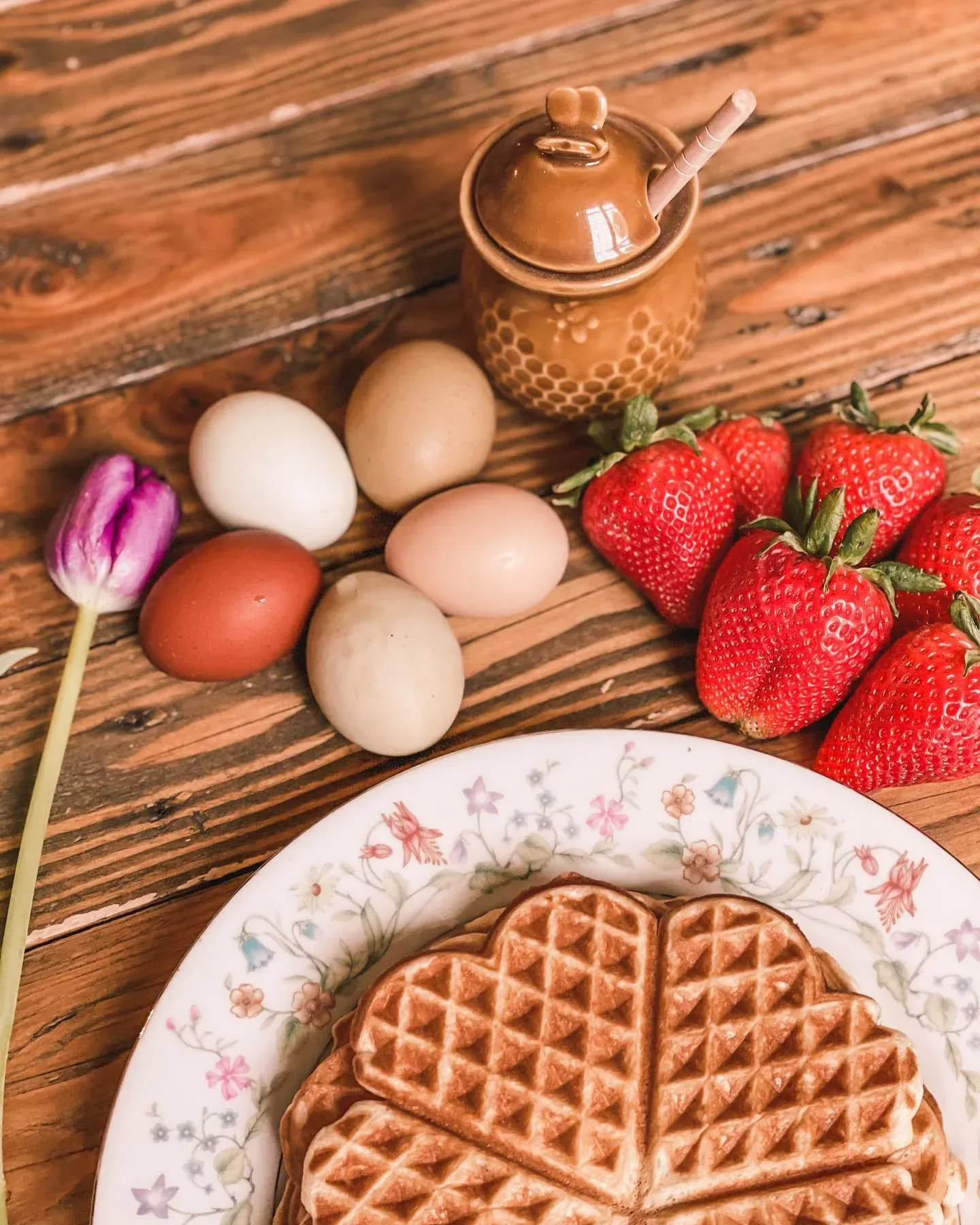 A waffle on a floral plate next to fresh eggs, strawberries, a small honey pot, and a single purple tulip on wood.