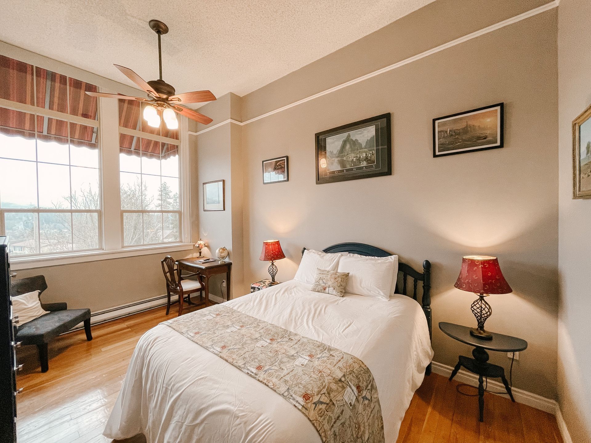 A cozy bedroom featuring a bed with white bedding, a desk by large windows, and two lamps on bedside tables.