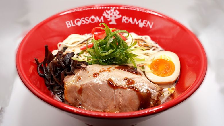 Red bowl of ramen with sliced pork, egg, green onions, and seaweed on a white table