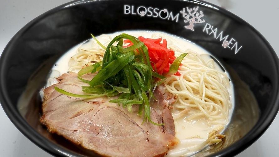 Steaming bowl of ramen with sliced pork, green onions, and red garnish in a black Blossom Ramen bowl