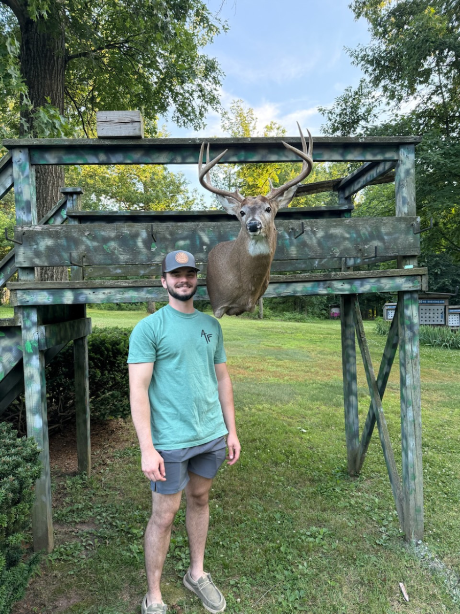 Man standing beside a mounted deer in a grassy outdoor hunting display