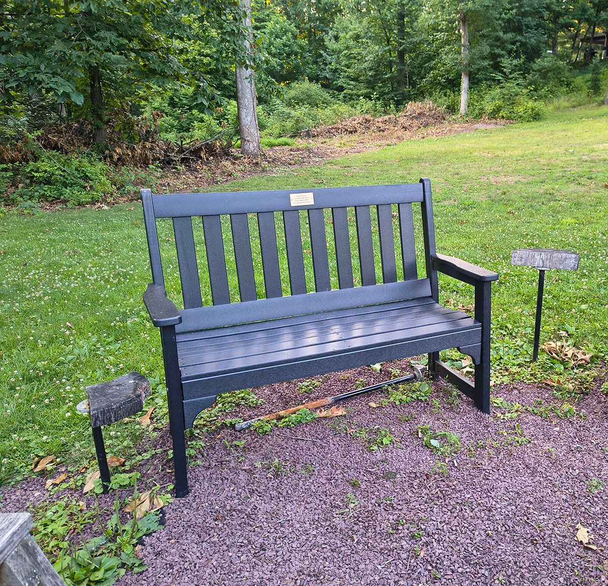 Black metal bench on gravel in a green park, with two small signs beside it.