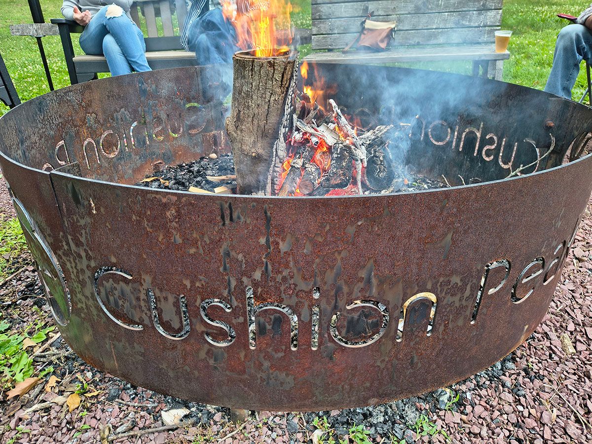 Rusty metal fire pit with a burning log and the words “Cushion Peak” cut into the side