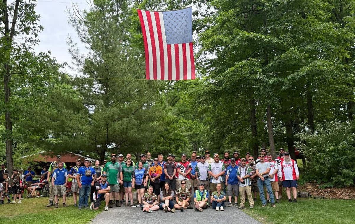 Large group posing outdoors beneath a hanging American flag in a wooded park.