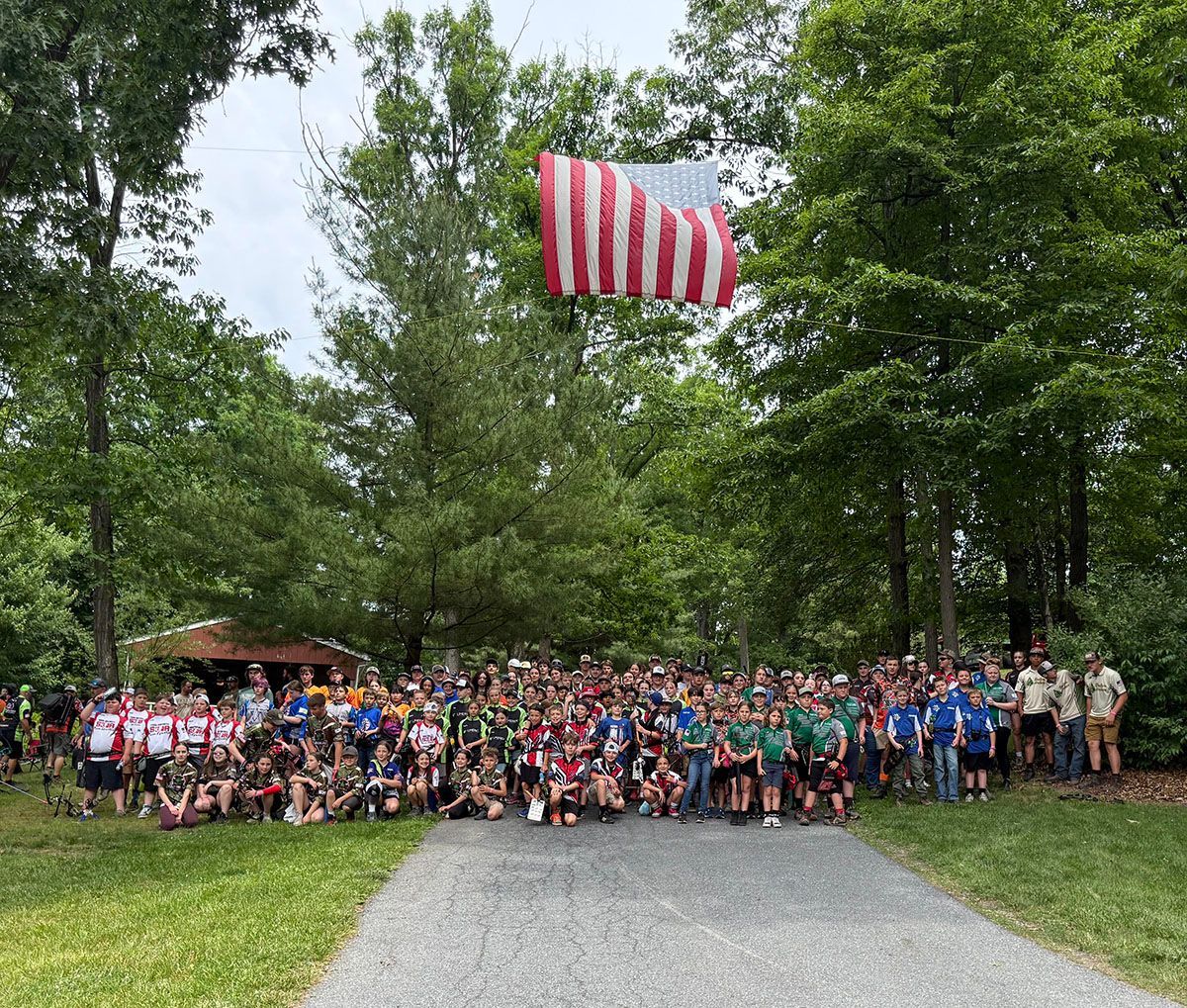 Large group posing outdoors on a path under trees, with a red-and-white striped flag above them.
